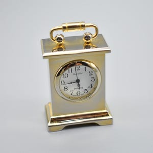 May include: A small, rectangular quartz clock with a gold-coloured frame and a white face. The clock has black numerals and hands, and a gold handle on top. The clock is approximately 10 cm tall.