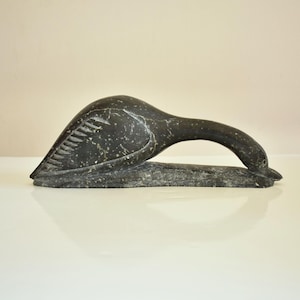 May include: A dark grey stone sculpture of a bird with a long neck and a curved handle. The bird's wing has carved lines. The sculpture is on a white surface.