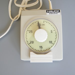 Op de afbeelding: Een vintage Hauck elektrische timer met een crèmekleurige wijzerplaat en een witte wijzerplaat. De timer heeft een maximum van 30 seconden en een witte knop. De timer is ondergebracht in een lichtgrijze rechthoekige behuizing met een wit snoer.