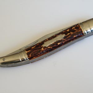 Peut inclure: Un couteau de poche pliant avec un cadre en métal fermé et un manche texturé aux motifs tachetés marron et noir. Le manche présente une incrustation métallique argentée allongée gravée du mot FISH KNIFE. Une petite boucle est fixée à une extrémité.