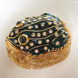 Peut inclure: Une boîte à bijoux décorative en forme de grenouille, dotée d'un corps émaillé vert foncé avec des bordures dorées et des cristaux transparents incrustés. La base est en métal doré poli avec une texture complexe rappelant des feuilles.