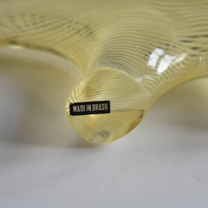 K&ouml;nnte beinhalten: Eine gelbe Glasskulptur mit einer wellenf&ouml;rmigen, organischen Form. Die Skulptur tr&auml;gt ein Etikett mit der Aufschrift "MADE IN BRASIL".