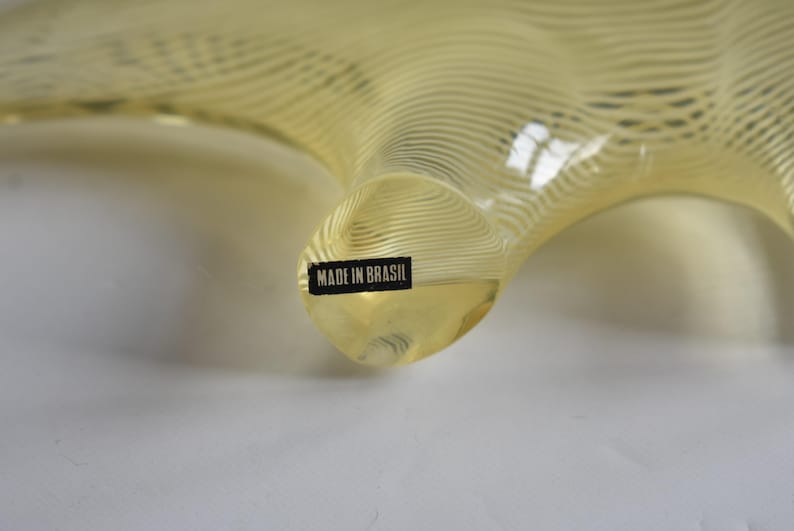 K&ouml;nnte beinhalten: Eine gelbe Glasskulptur mit einer wellenf&ouml;rmigen, organischen Form. Die Skulptur tr&auml;gt ein Etikett mit der Aufschrift "MADE IN BRASIL".
