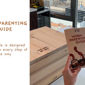 Op de afbeelding: Een boek getiteld "Worm Parenting Guide" wordt voor een houten kist gehouden. De omslag van het boek bevat illustraties van wormen en de tekst "Worm Parenting Guide". De tekst "The Box of Life" staat op de kist.