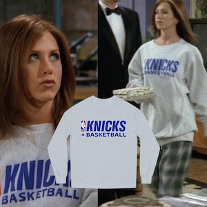 May include: A light gray crewneck sweatshirt featuring the blue text KNICKS BASKETBALL and the NBA logo on the chest. The long-sleeve pullover has a ribbed collar, cuffs, and hem, with a Champion brand logo patch on the left sleeve near the cuff.