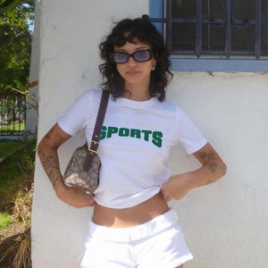 May include: A white t-shirt with a green "SPORTS" graphic. The person is wearing white shorts and sunglasses.