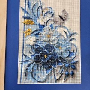 Op de afbeelding: Een ingelijst borduurwerk met kruissteek, met een boeket blauwe, witte en gele bloemen met een vlinder. De bloemen zijn in een vaas gerangschikt en de achtergrond is lichtblauw.