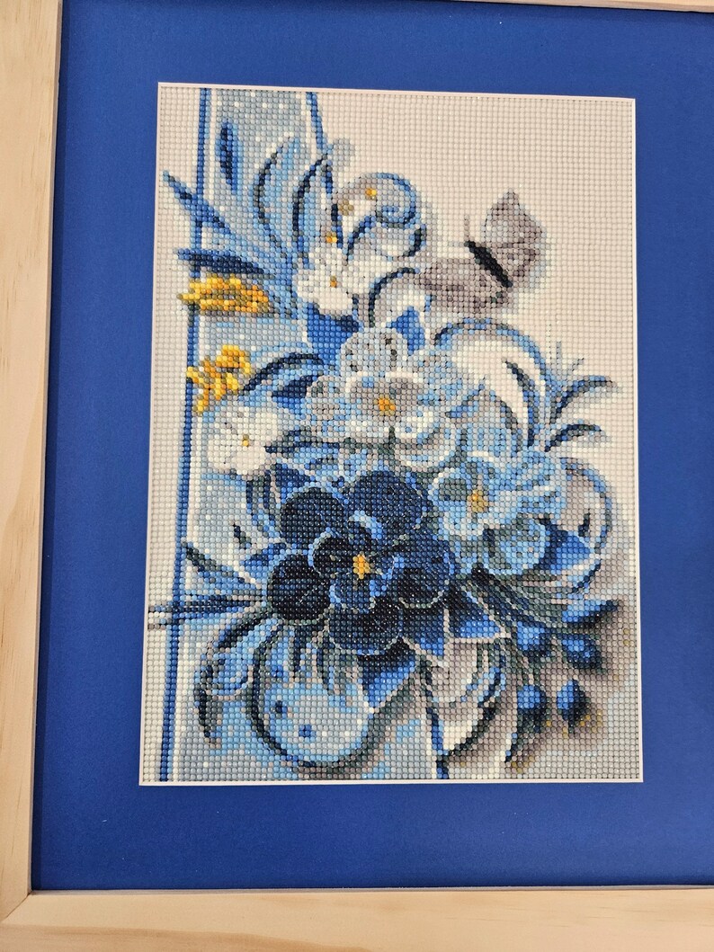 Op de afbeelding: Een ingelijst borduurwerk met kruissteek, met een boeket blauwe, witte en gele bloemen met een vlinder. De bloemen zijn in een vaas gerangschikt en de achtergrond is lichtblauw.