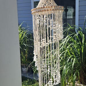 May include: A hanging seashell chandelier with a tiered design. The top section is made of small, light-colored shells, while the lower portion features strands of white shells. The chandelier hangs outdoors.