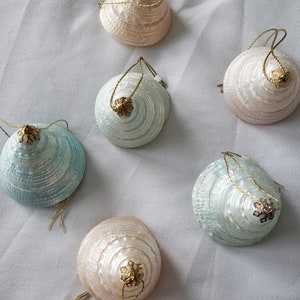 May include: Six iridescent Christmas ornaments shaped like seashells with gold accents and gold string hangers. The ornaments are in shades of pink, blue, and white.