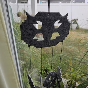 May include: A dark gray wind chime with a textured surface, featuring a stylized owl design with cut-out shapes. The chime hangs from black cords, with several decorative pieces suspended below. The background includes a window, greenery, and houses.
