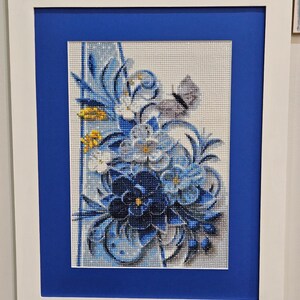 Op de afbeelding: Een ingelijst diamanten schilderij met een blauw en wit bloemenmotief met een vlinder. De bloemen zijn in een boeket gerangschikt met een blauwe achtergrond.