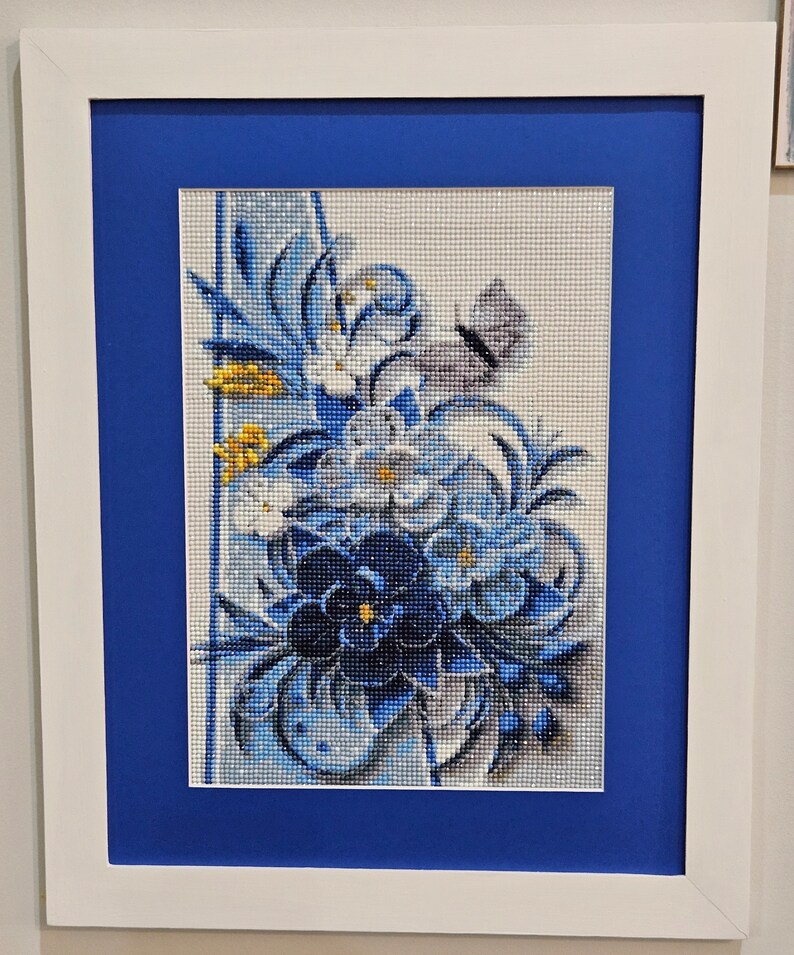 Op de afbeelding: Een ingelijst diamanten schilderij met een blauw en wit bloemenmotief met een vlinder. De bloemen zijn in een boeket gerangschikt met een blauwe achtergrond.