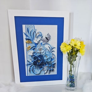 Op de afbeelding: Een ingelijst borduurwerk met kruissteek, met een blauw en wit bloemenmotief met een vlinder. Het borduurwerk is in een wit frame met een blauwe passe-partout.
