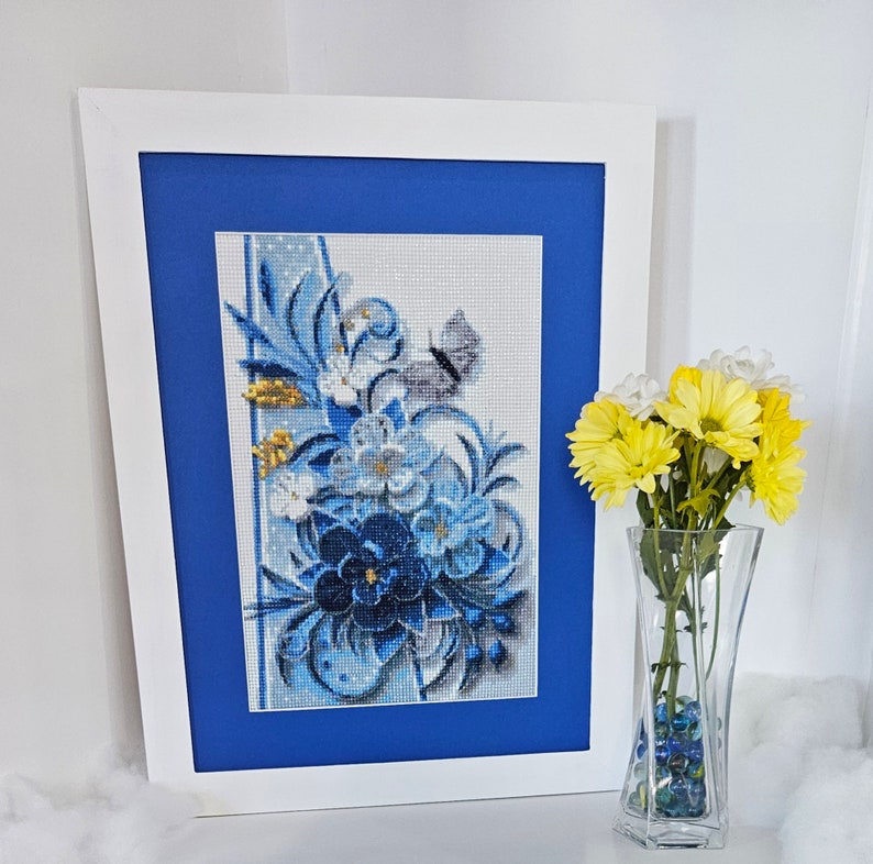 Op de afbeelding: Een ingelijst borduurwerk met kruissteek, met een blauw en wit bloemenmotief met een vlinder. Het borduurwerk is in een wit frame met een blauwe passe-partout.