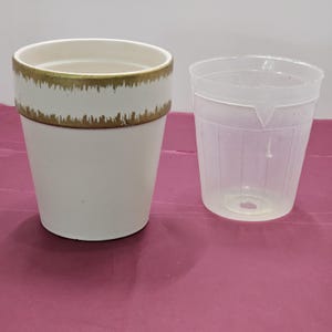 May include: A white ceramic plant pot with a gold-painted rim and a clear plastic pot. The ceramic pot has a cylindrical shape. The clear plastic pot is ribbed and has a tapered shape.