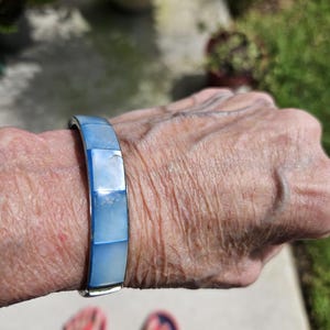 May include: A silver-toned bracelet with rectangular, light blue iridescent segments. The bracelet is worn on a wrist, with a blurred background of greenery and a person's feet in red sandals.