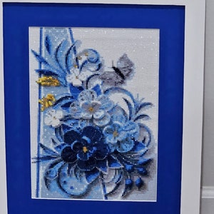 Op de afbeelding: Een ingelijst diamanten schilderij met een blauw en wit bloemenontwerp met een vlinder. Het schilderij is op een witte achtergrond en ingelijst met een blauwe passe-partout en een wit frame.