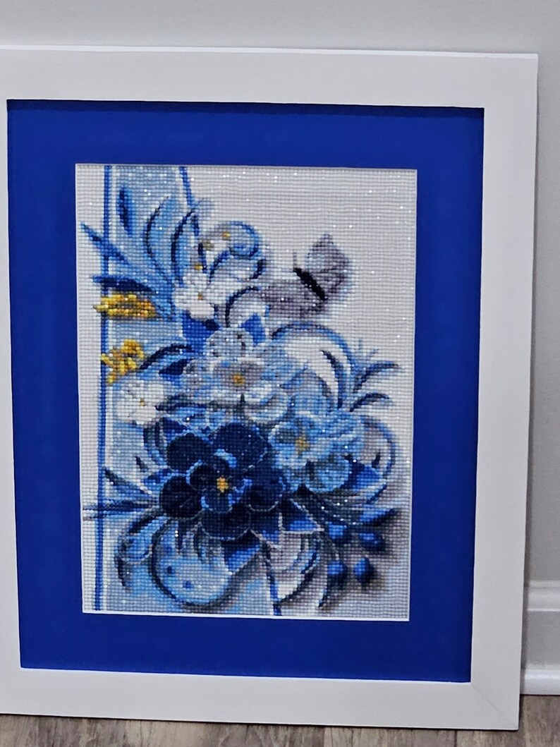 Op de afbeelding: Een ingelijst diamanten schilderij met een blauw en wit bloemenontwerp met een vlinder. Het schilderij is op een witte achtergrond en ingelijst met een blauwe passe-partout en een wit frame.