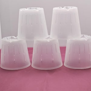 May include: A collection of five translucent white plastic plant pot covers. Each cover is cone-shaped with vertical slits and a small opening at the top. The covers are stacked and arranged on a pink surface.