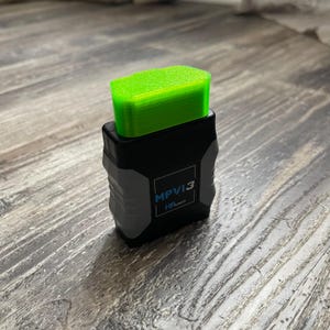 May include: A black and gray MPVI3 device with a bright green top. The device has the text "MPVI3" and "HP Tuners" on the front. The device is sitting on a wooden surface.