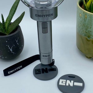 Könnte beinhalten: Ein silberner ENHYPEN-Lichtstick mit einer durchsichtigen Plastikkugel oben. Der Lichtstick hat ein schwarzes Band mit dem Wort "ENHYPEN" darauf gedruckt. Es gibt zwei graue runde Scheiben mit dem Wort "EN" darauf gedruckt.