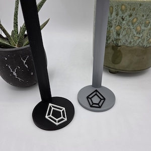 May include: Two geometric design stands. One is black with a white pentagon design, and the other is gray with a black pentagon design. The stands have a circular base and a vertical support.