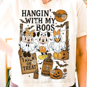 May include: Cream-colored t-shirt with a Halloween-themed graphic. The design features Mickey, Minnie, and Daisy Duck dressed as ghosts, along with pumpkins, bats, and the text "HANGIN' WITH MY BOOS" and "TRICK OR TREAT".