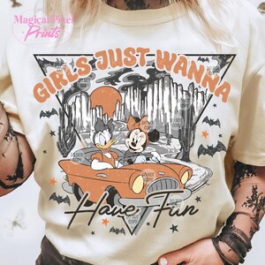 May include: A white t-shirt with a vintage-style graphic featuring two cartoon characters driving a red car. The text "Girls Just Wanna Have Fun" is written in a retro font.