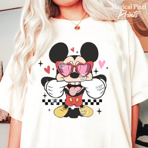 Pu&ograve; includere: Maglietta bianca con un'immagine di cartone animato di Topolino che indossa occhiali da sole a forma di cuore e un papillon rosso. Il design include cuori rossi, stelle e un motivo a scacchi.