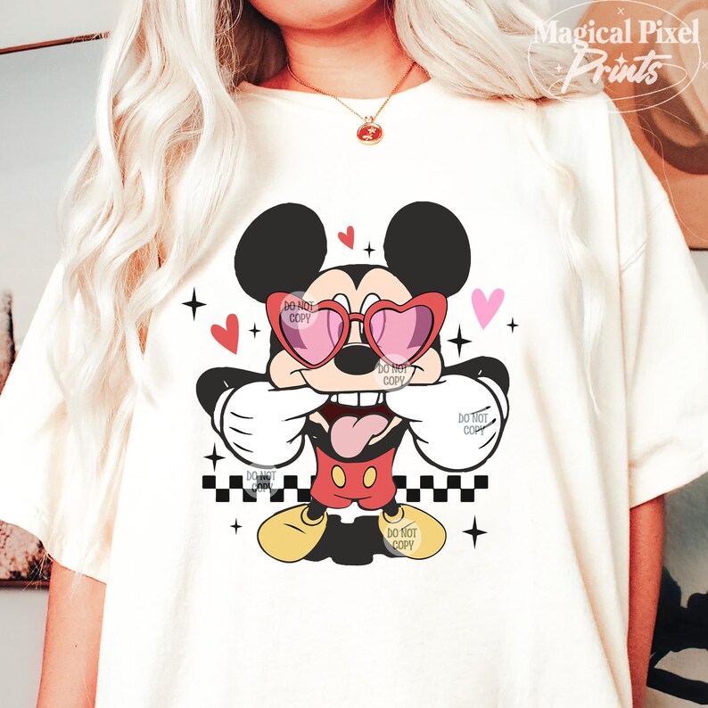 Pu&ograve; includere: Maglietta bianca con un'immagine di cartone animato di Topolino che indossa occhiali da sole a forma di cuore e un papillon rosso. Il design include cuori rossi, stelle e un motivo a scacchi.