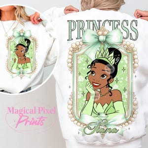 May include: White sweatshirt with a green and gold princess design. The design features a cartoon princess with a crown, a green dress, and a frog. The text "Princess" is above the design, and the text "Tiana" is below the design.