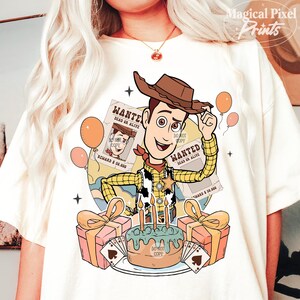 May include: A white t-shirt with a cartoon image of Woody from Toy Story. Woody is wearing his signature cowboy hat and vest. He is holding a wanted poster and standing in front of a birthday cake with candles. The text on the wanted posters reads "WANTED DEAD OR ALIVE".