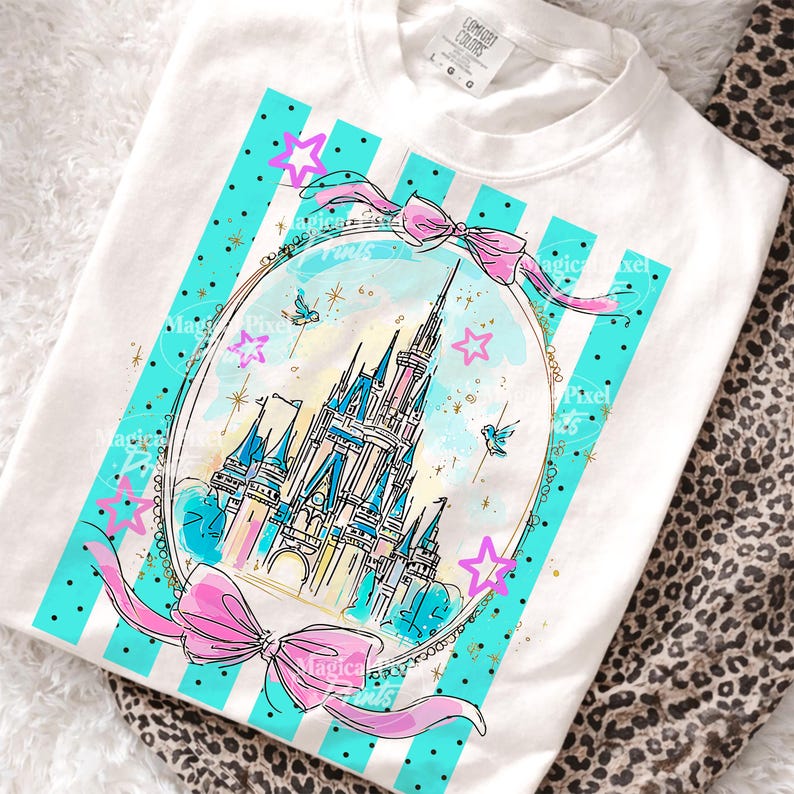 May include: White t-shirt with a watercolour illustration of a castle inside a decorative frame. The design includes turquoise stripes, pink bows, stars, and small birds. The text "Magical Pixels" is visible on the design.