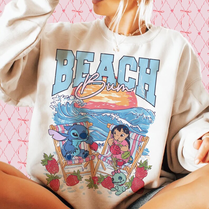 Puede incluir: Sudadera crema con un gr&aacute;fico de tem&aacute;tica playera. El dise&ntilde;o presenta las palabras "BEACH Bum" en azul y rosa, con los personajes de dibujos animados Stitch y Lilo en sillas de playa, rodeados de fresas.