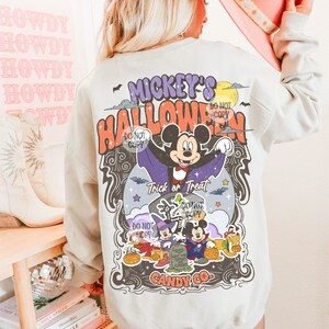 Może przedstawiać: Jasnoszara bluza z kolorową grafiką Halloween. Projekt przedstawia Myszka Miki przebranego za wampira, z napisami "Mickey's Halloween" i "Trick or Treat". Bluza ma r&oacute;wnież logo "Candy Co.".