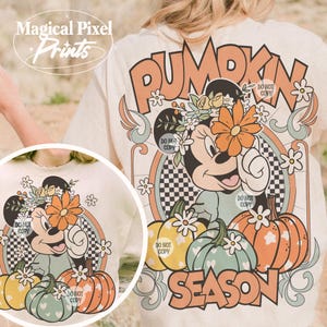 May include: A beige t-shirt with a vintage-style graphic featuring a cartoon mouse wearing a flower crown and holding a large orange flower. The graphic also includes pumpkins, flowers, and the text "Pumpkin Season".