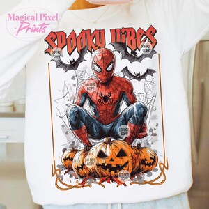 K&ouml;nnte beinhalten: Wei&szlig;es Langarmshirt mit einem Spiderman-Motiv und dem Text "Spooky Vibes". Das Design umfasst Flederm&auml;use, Spinnweben und geschnitzte K&uuml;rbisse. Das Shirt hat ein Halloween-Thema.