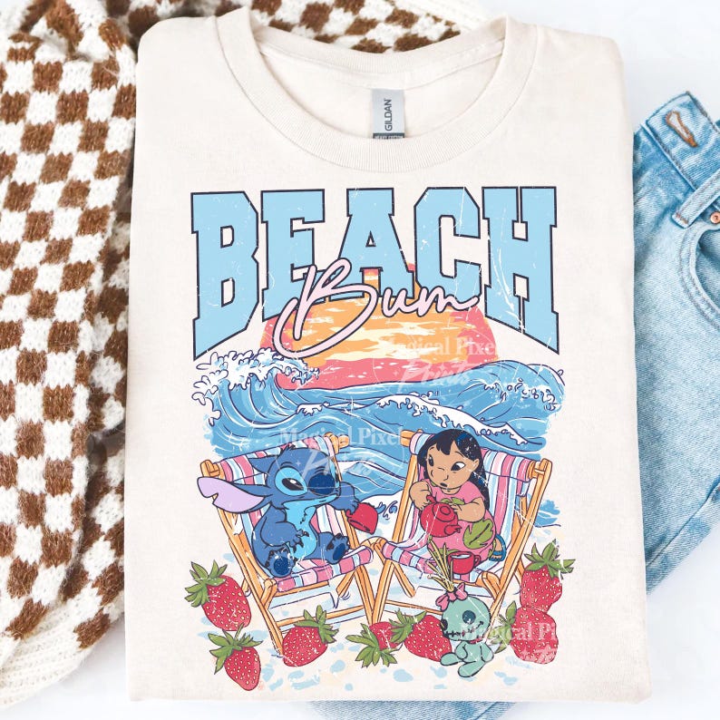 Puede incluir: Camiseta color crema con un dise&ntilde;o gr&aacute;fico con las palabras "BEACH Bum" en azul y rosa. El dise&ntilde;o incluye personajes de dibujos animados, sillas de playa, olas y fresas. La camiseta est&aacute; doblada.