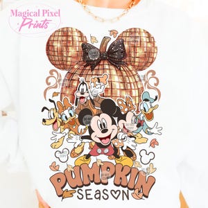 K&ouml;nnte beinhalten: Ein wei&szlig;es T-Shirt mit einem K&uuml;rbis-Design, das Mickey Mouse und seine Freunde zeigt. Der K&uuml;rbis ist aus goldenem Glitzer und hat einen Discokugel-Effekt. Der Text "Pumpkin Season" ist in orangefarbenen und braunen Buchstaben mit einem Herzen im Wort "Season" geschrieben.