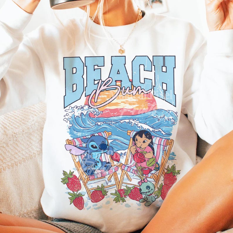 Puede incluir: Sudadera blanca con un dise&ntilde;o gr&aacute;fico. El dise&ntilde;o presenta el texto "BEACH Bum" en azul y rosa, personajes de dibujos animados, sillas de playa, olas y fresas, creando un ambiente veraniego.