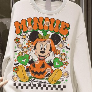 May include: White sweatshirt featuring a Halloween-themed Minnie Mouse design. The design includes the word "MINNIE" in orange and green, Minnie Mouse in a pumpkin costume, flowers, hearts, and the text "Minnie Mouse xoxo".