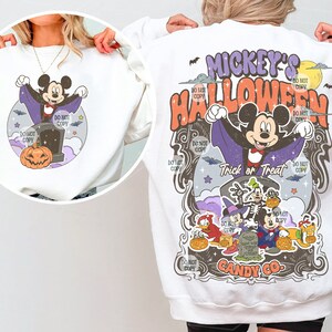Może przedstawiać: Biała bluza z nadrukiem o tematyce Halloween. Projekt zawiera Myszkę Miki przebraną za wampira, dynię i napis "Mickey's Halloween". Widoczne są r&oacute;wnież inne postacie. Widoczne są r&oacute;wnież słowa "Trick or Treat" i "Candy Co."