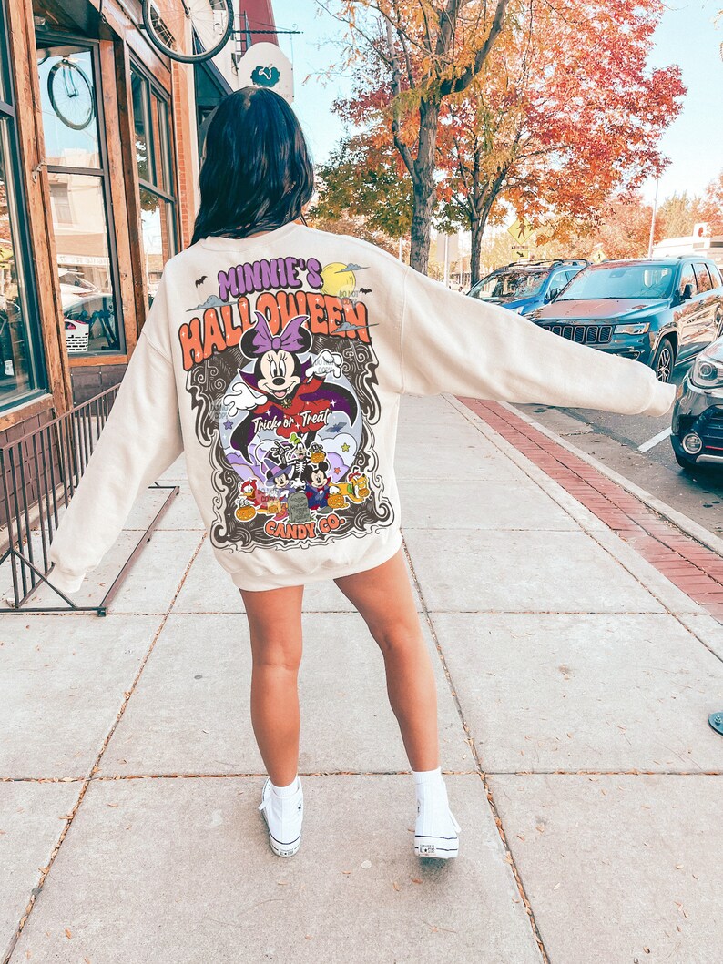May include: A cream colored sweatshirt with a graphic print of Minnie Mouse dressed as a vampire. The graphic says "Minnie's Halloween" and "Trick or Treat" with a candy corn design.
