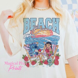 Puede incluir: Camiseta color crema con un dise&ntilde;o de playa. El dise&ntilde;o presenta las palabras "BEACH Bum" sobre una escena con los personajes de dibujos animados Stitch y Lilo, olas, una puesta de sol y fresas.