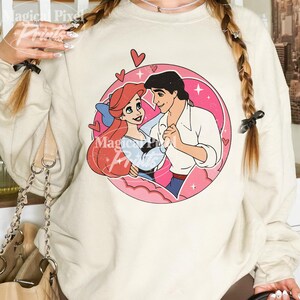 May include: A cream-colored sweatshirt with a graphic print of Ariel and Prince Eric from The Little Mermaid. The graphic is in a circle with pink hearts and clouds. The text "Magical Prints" is visible in the bottom left corner of the image.