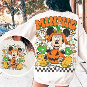 May include: Cream-colored sweatshirt featuring a Halloween-themed Minnie Mouse design. The graphic includes Minnie Mouse dressed as a pumpkin, surrounded by flowers, hearts, and the word "MINNIE" in orange and green. A black and white checkered pattern is at the bottom.