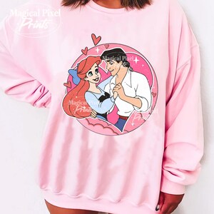 May include: Pink sweatshirt with a graphic print of Ariel and Prince Eric from The Little Mermaid. The graphic is in a circle with hearts and stars around it.