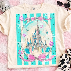 May include: Cream-coloured t-shirt with a fairytale castle design within a teal and pink frame. The design includes a castle, stars, and bows. The shirt has a classic crew neck and short sleeves, suitable for casual wear.