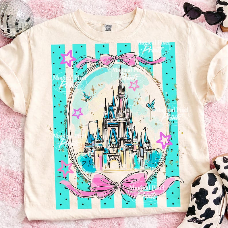 May include: Cream-coloured t-shirt with a fairytale castle design within a teal and pink frame. The design includes a castle, stars, and bows. The shirt has a classic crew neck and short sleeves, suitable for casual wear.